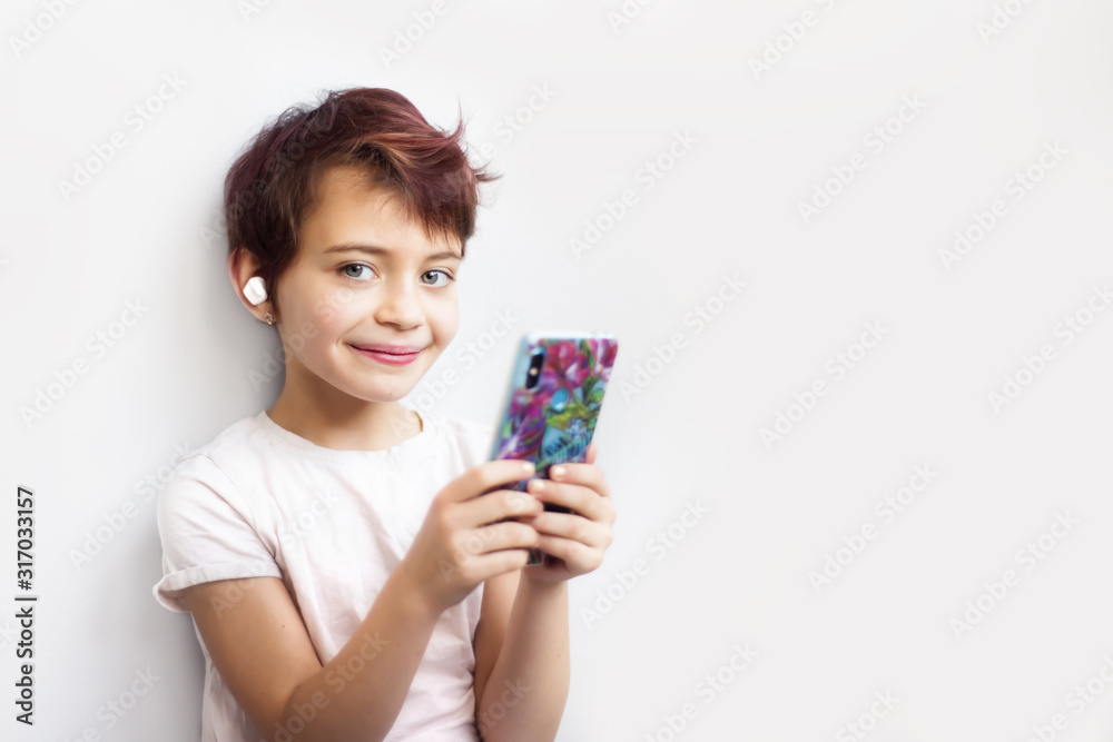 Horizontal portrait little 7 years old short haired schoolgirl with pink hair looking in camera and listening music on mobile phone with white wireless headphones. Gadget addicted children. Horizontal portrait little 7 years old short haired schoolgirl with pink hair looking in camera and listening music on mobile phone with white wireless headphones. Gadget addicted children.