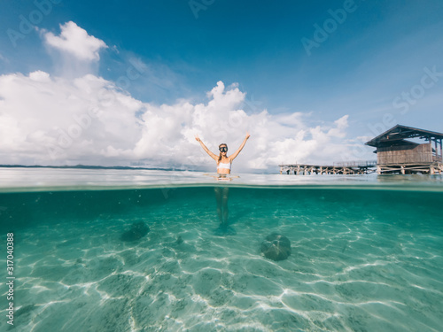 Split shot portrait of carefree female scuba diver in mask for snorkeling submerged in crystal water with pier on background, woman enjoying summer vacation at tropical island with tranquil waterline