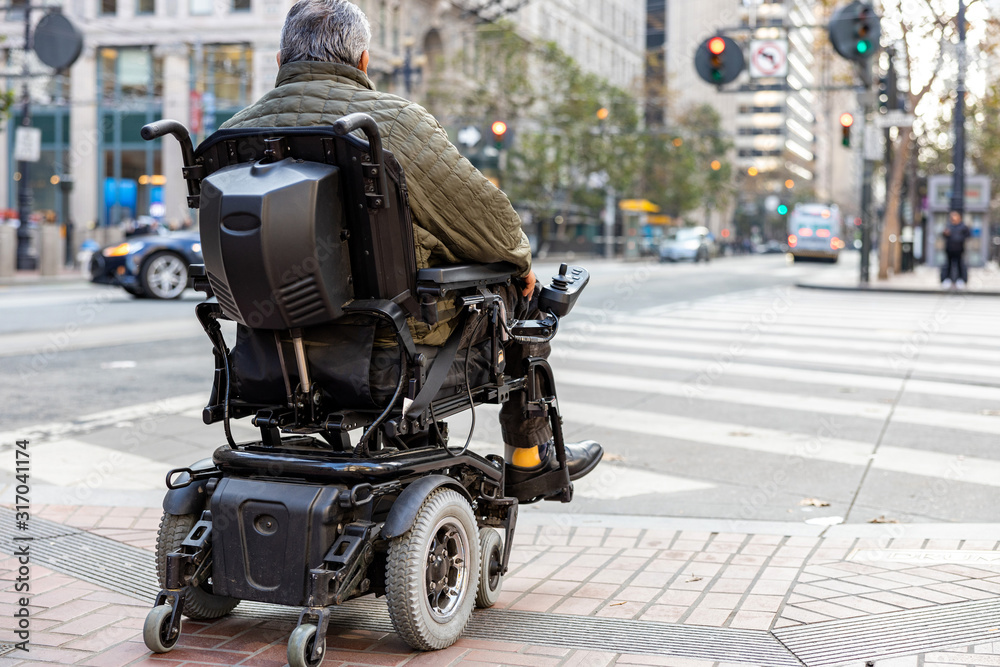 An elderly disabled person on an electric wheelchair on a city street in front of a pedestrian crossing. The concept of modern technology for people with disabilities. - obrazy, fototapety, plakaty 