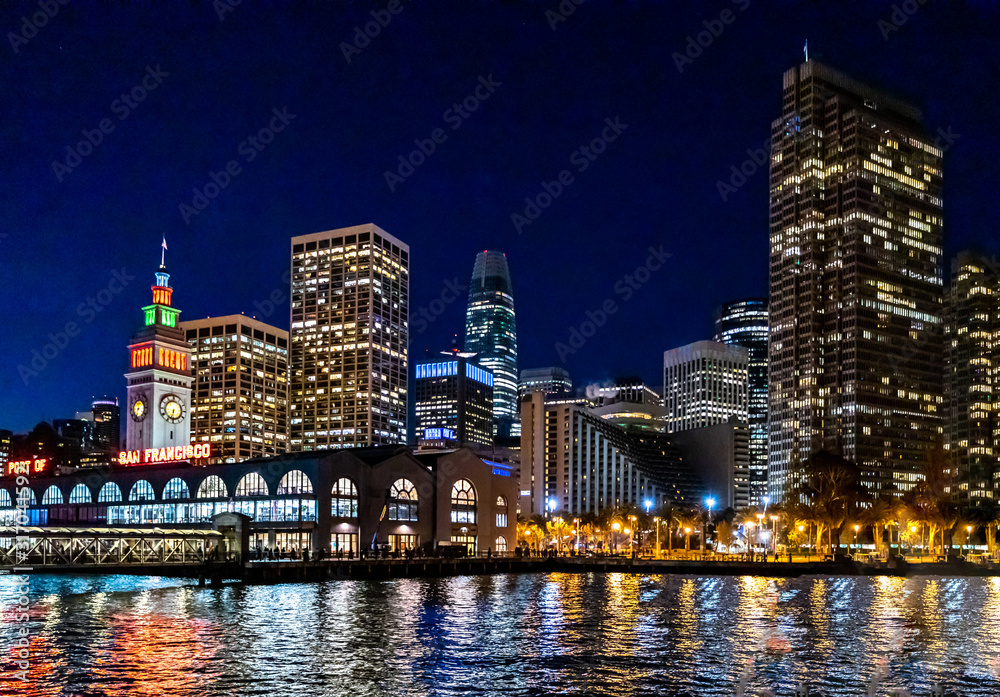 Panoramic view of the port of San Francisco, illuminated at night, against the backdrop of impressive skyscrapers with light and reflection in the water, view from the side of the San Francisco Bay.