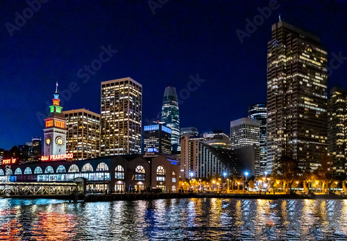 Panoramic view of the port of San Francisco, illuminated at night, against the backdrop of impressive skyscrapers with light and reflection in the water, view from the side of the San Francisco Bay.