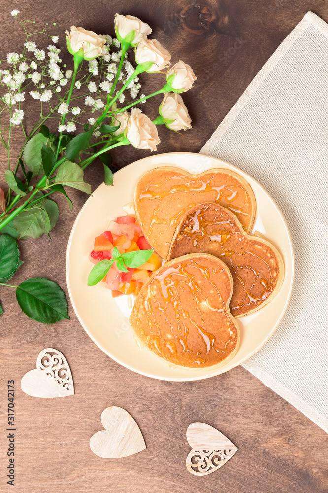 Fototapeta premium Homemade heart shaped pancakes with fruit chutney salad and honey on a beige plate on a wooden table. Rustic style breakfast or brunch for Valentine's Day. Top view, vertical orientation