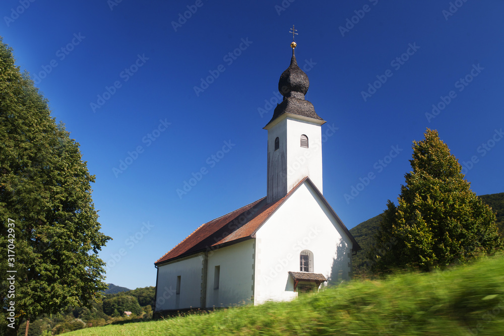 Little white  church in Slovenian countryside among lush green summer landscape and blue sky