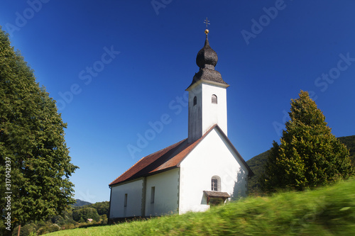 Little white church in Slovenian countryside among lush green summer landscape and blue sky