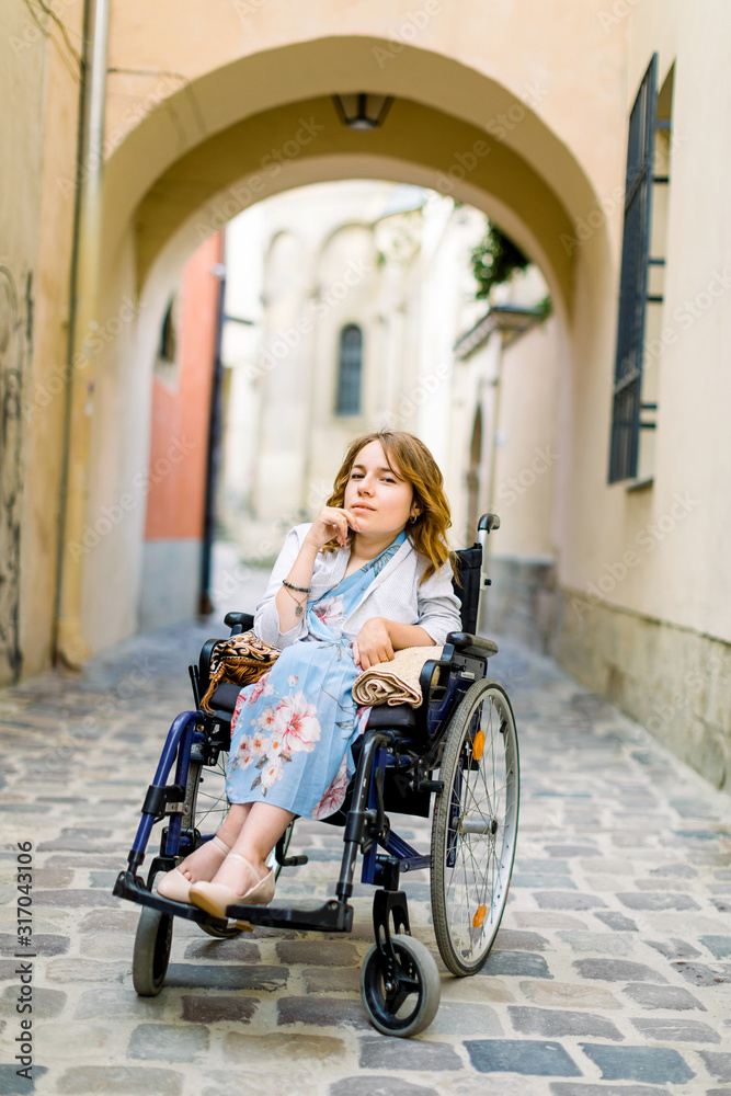 Portrait of young blond-haired woman sitting in wheelchair and looking at camera, walking outdoors in old city center, building arch on the background