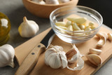 Bowl of garlic slices, bulbs, oil, knife, board on grey wooden background. Closeup