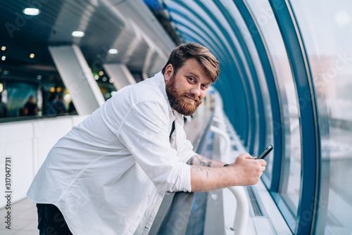 Portrait of handsome hipster guy in casual outfit holding modern cellphone gadget for mobility and chatting, bearded man with cellular device looking at camera while resting in public place