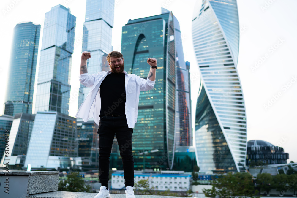 Full length portrait of overjoyed hipster guy with raised hands smiling at camera at urban setting celebrating completed sightseeing in metropolis, happy man posing at skyscrapers background Full length portrait of overjoyed hipster guy with raised hands smiling at camera at urban setting celebrating completed sightseeing in metropolis, happy man posing at skyscrapers background