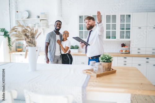 Принти на полотні Confident estate agent showing kitchen to African American pleased hugging coupl