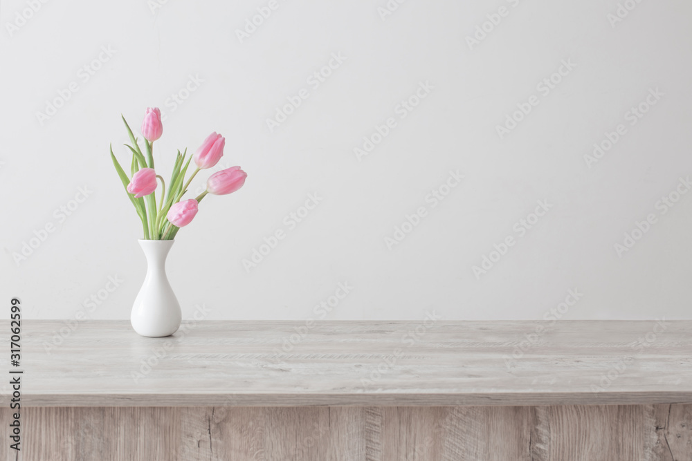 pink tulips in white ceramic vase on wooden table on background white wall