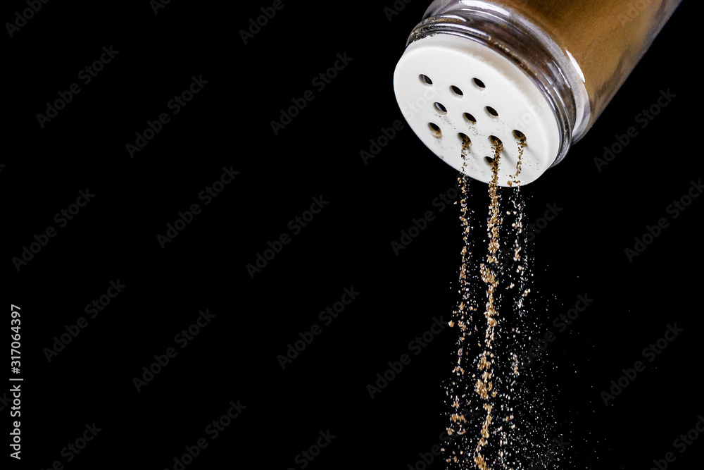 Black ground pepper falls out of the pepper shaker on a black isolated background. The seasoning process stop in time. Glass salt shaker with white strainer.