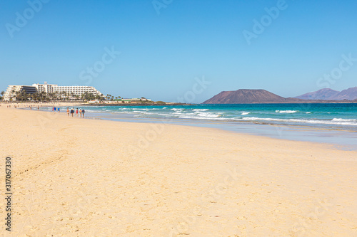 Fototapeta Naklejka Na Ścianę i Meble -  Beach hotel on Fuerteventura canary islands with the beach in front and the sea with blue azure aquamarine turquoise colors and a vulcano in the background blue sky