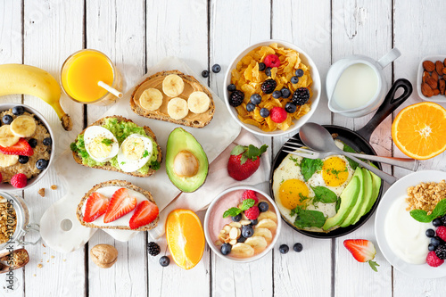 Healthy breakfast table scene with fruits, yogurts, oatmeal, cereal, smoothie bowl, nutritious toasts and egg skillet. Top view over a white wood background.