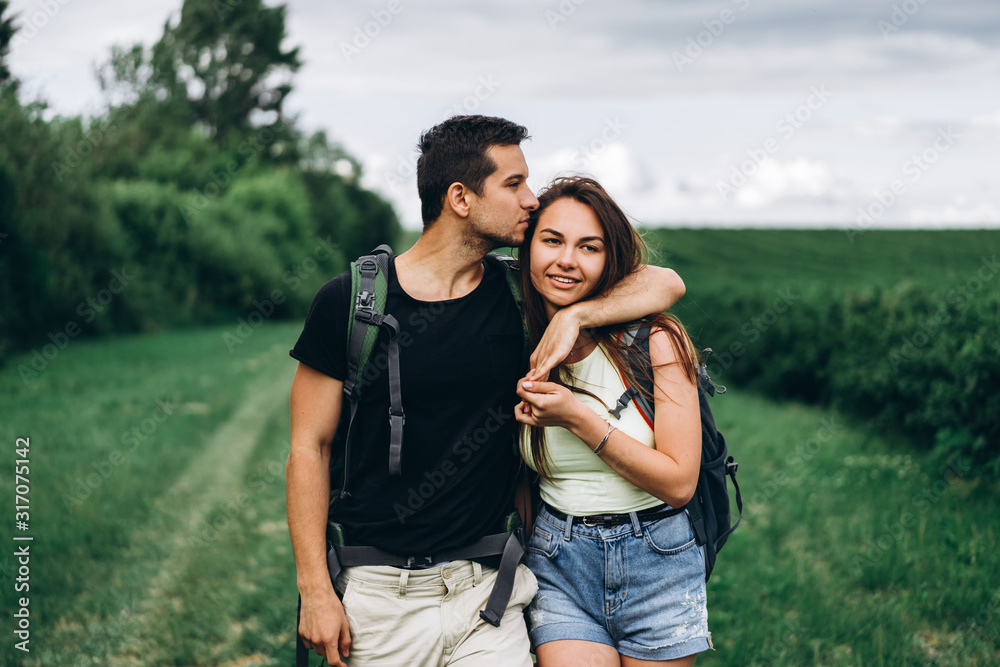 Portrait of happy young couple with backpacks on the field in spring. Man and woman walking in currant plantations Portrait of happy young couple with backpacks on the field in spring. Man and woman walking in currant plantations