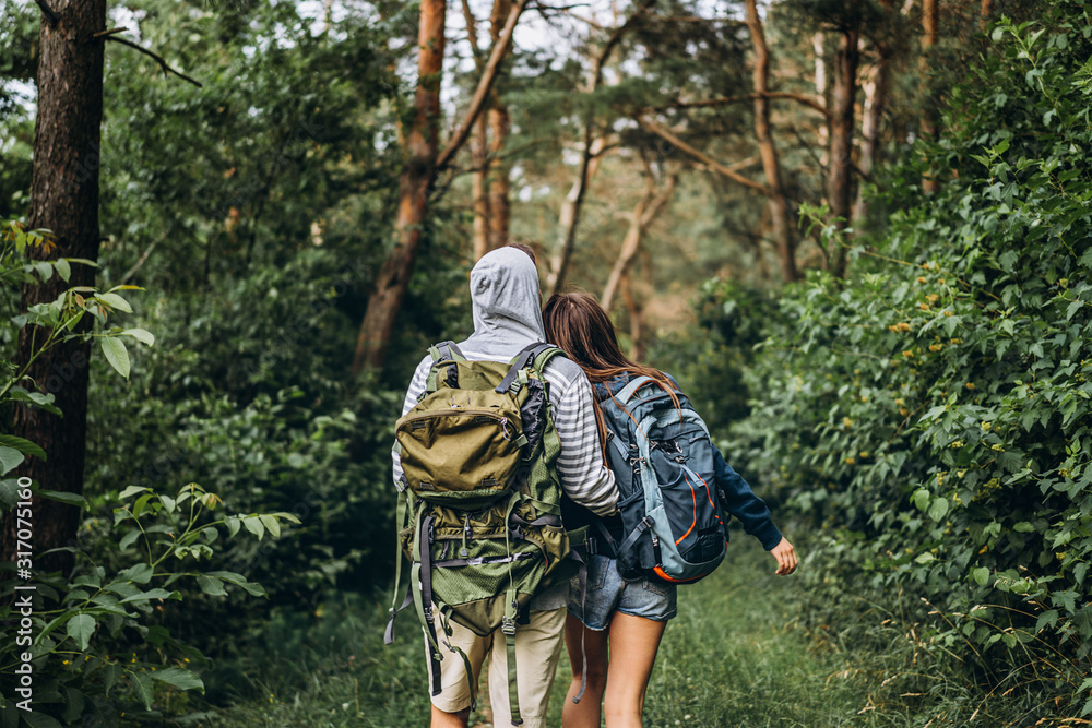 Back view of young couple with backpacks on their backs in the forest. Loving man hugging his beautiful girlfriend on a hike in the woods