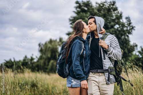 Young loving couple with backpacks walking and gently hugging on the field and trees background. Love Story