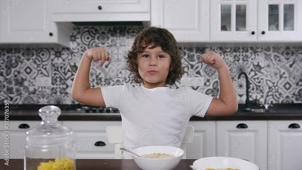 Smiling small boy sitting at kitchen table and shows strength in his hands after his breakfast of delicious healthy meal of corn flakes and milk. Dolly shot Smiling small boy sitting at kitchen table and shows strength in his hands after his breakfast of delicious healthy meal of corn flakes and milk. Dolly shot