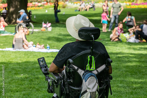 rear view of disabled man with backpack wearing sun hat while sitting by camera on black wheelchair over grassy lawn in park