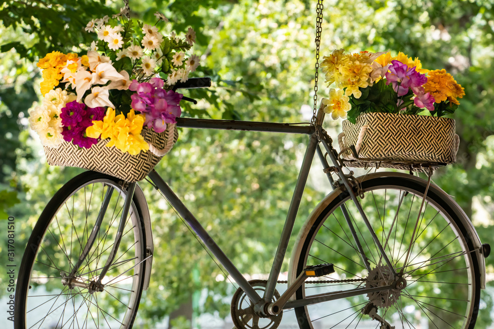 Multicolored fresh flower bouquets in baskets on metallic bicycle hanging with metallic chain against tree backdrop on sunny day at garden