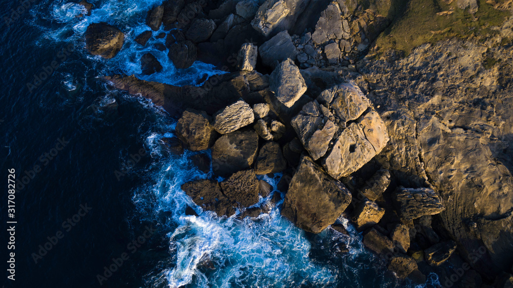 Top view of the coast. In the image we can see the blue sea with some rocks on a colorful sunset.