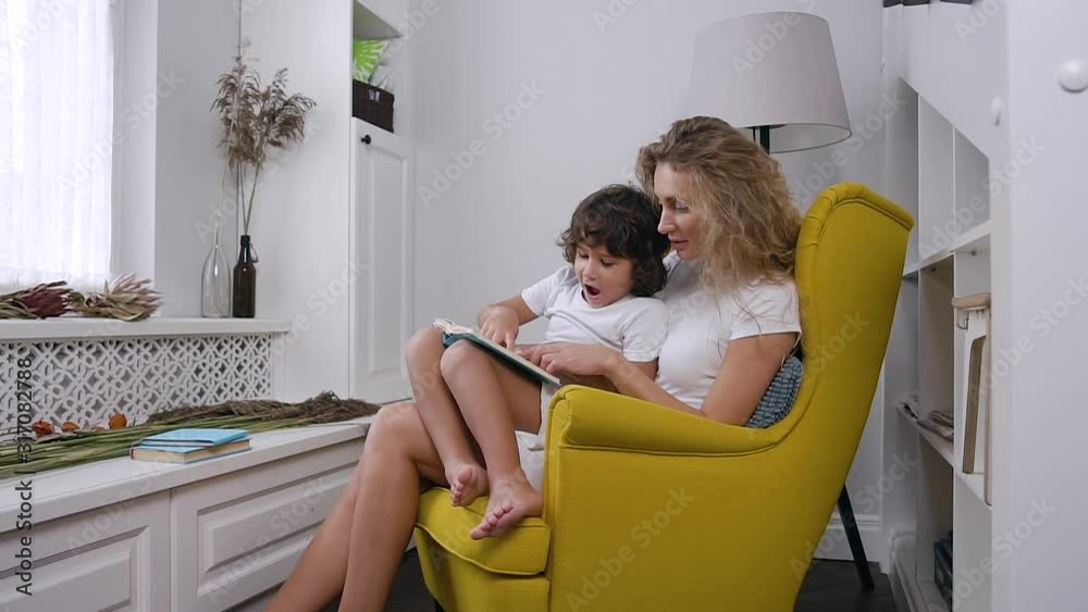 A young lovely mom reads child book to her little son while sitting in comfortable yellow armchair near window at cozy home. A young lovely mom reads child book to her little son while sitting in comfortable yellow armchair near window at cozy home.