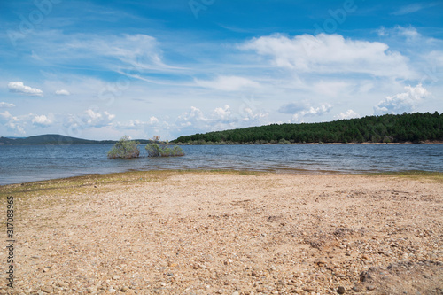 Shore of a huge natural lake  surrounded by trees  under a beautiful blue sky with clouds.