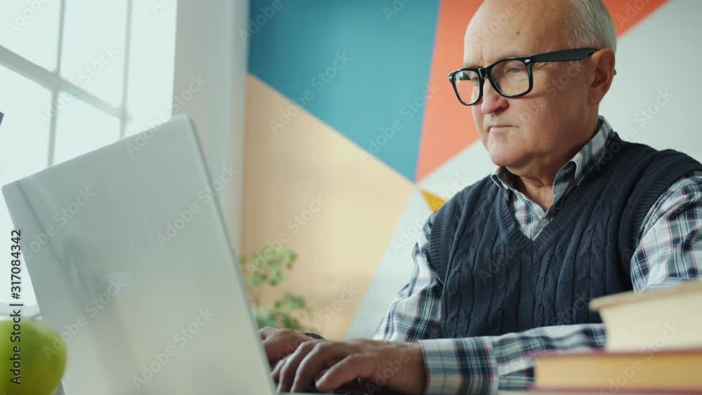 Slow motion of retired man using laptop computer typing sitting alone at desk in apartment. Contemporary technology, retirement and modern lifestyle concept. Slow motion of retired man using laptop computer typing sitting alone at desk in apartment. Contemporary technology, retirement and modern lifestyle concept.