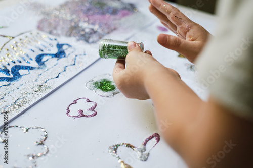 The kid's hands holding the container of green sparkles and sprinkling glitter for creating artwork