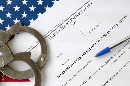 District court warrant for the arrest of a witness in a civil action papers with handcuffs and blue pen on United States flag. Permission to witness arrest