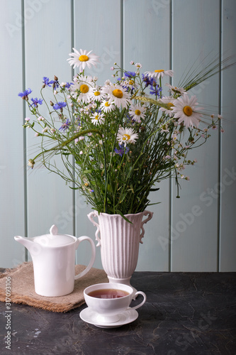 White cup with tea, white kettle and white vase with wildflowers cornflowers and daisies.