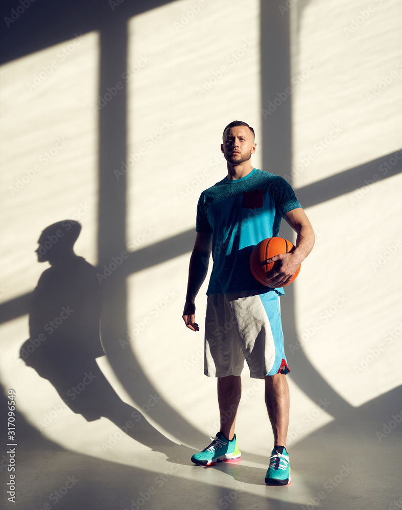 Young athletic man, basketball player holding ball and standing near wall with shadows from window