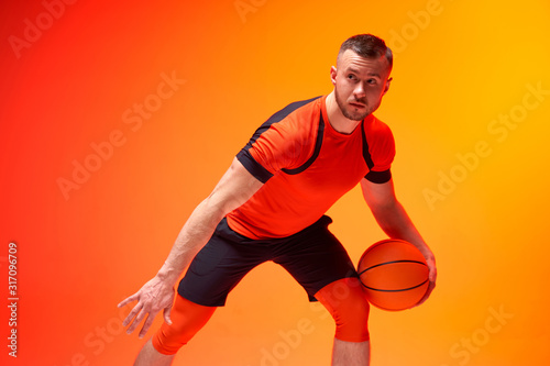 Young athletic man, basketball player standing with ball in defence position on orange and red background