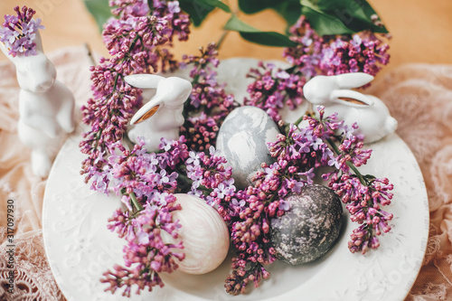 Happy Easter. Stylish Easter eggs on vintage plate, white bunnies and lilac flowers on fabric on wooden table. Rural composition of colorful natural dyed easter eggs and spring flowers