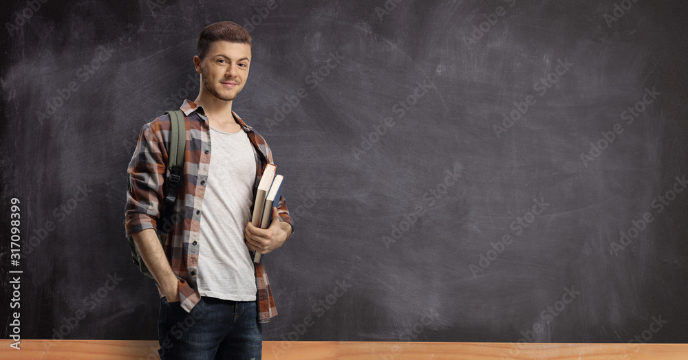 Male student in front of a blackboard holding books Male student in front of a blackboard holding books