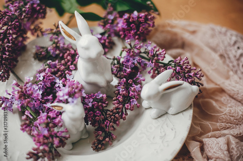Happy Easter. Stylish Easter white bunnies on vintage plate and lilac flowers on rural fabric. Ceramic rabbits decorations and spring flowers. Space for text. Holiday decor
