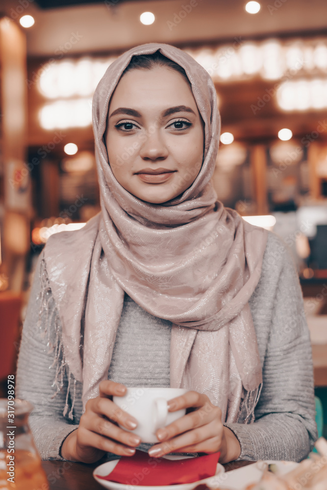 beautiful muslim woman in a headscarf gently looks at the camera with a cup of coffee in a cafe beautiful muslim woman in a headscarf gently looks at the camera with a cup of coffee in a cafe