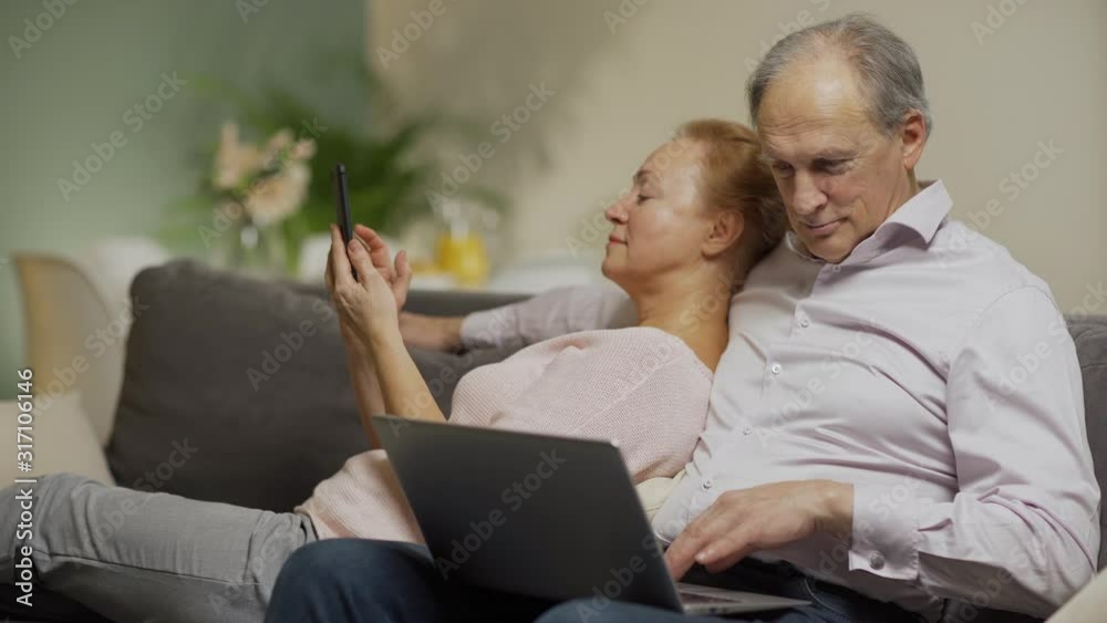 Contemporary senior couple spending leisure time at home. Man sitting on sofa and using laptop computer, wife leaning on him and text messaging on smartphone Contemporary senior couple spending leisure time at home. Man sitting on sofa and using laptop computer, wife leaning on him and text messaging on smartphone