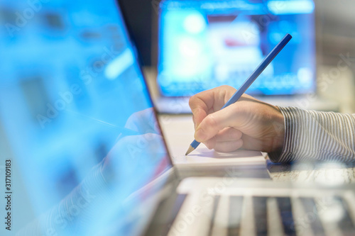 Closeup of hand arm of woman writing working  with a pen on a document close to her computer laptop pc in her home office on her workplace, desktop career screen notebook homework business blurred