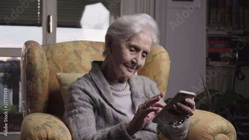 94 years old skilled lady portrait using smartphone technologies to communicate easily from her living room, senior woman sitting in yellow armchair holding phones and texting to family. 