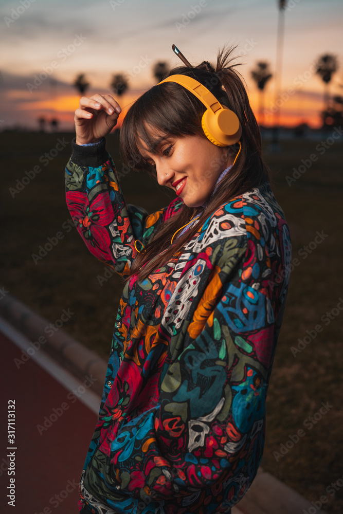 Close Up shot of Young handsome caucasian brown hair woman listening music with yellow headphones in a city park during amazing sunset. Music concept.