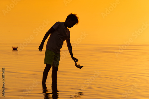 Little boy launch paper ship on the water. Beautiful summer sunset. Paper boat. Origami.