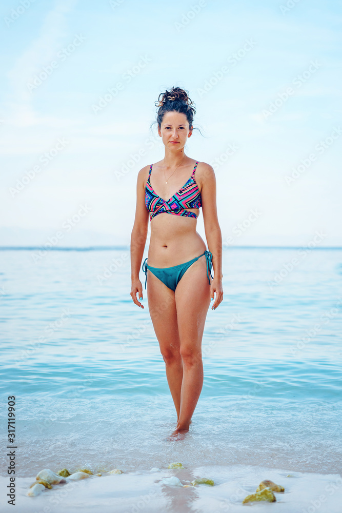 Full length portrait of young caucasian woman girl female standing on the beach in summer day wearing bikini swimsuit in front of the sea or ocean looking to the camera