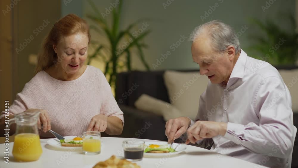Cheerful senior couple sitting at table at home and talking while having eggs for breakfast together, medium shot Cheerful senior couple sitting at table at home and talking while having eggs for breakfast together, medium shot