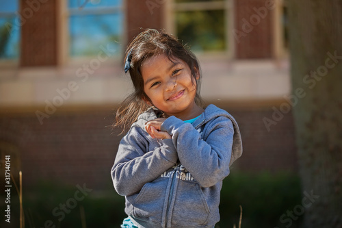 Cute little girl holding her hands together who is expressing love, happiness or grattitude, with a school building made out of bricks in background.