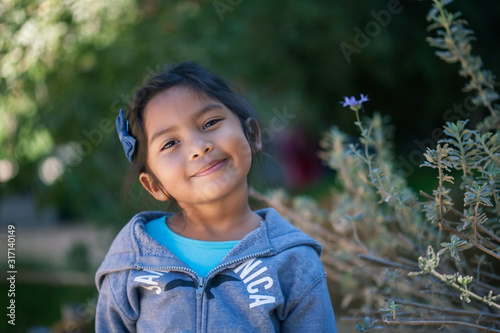 Hispanic young girl contemplating her choices or making up her mind, who is standing in outdoors setting with green plants.