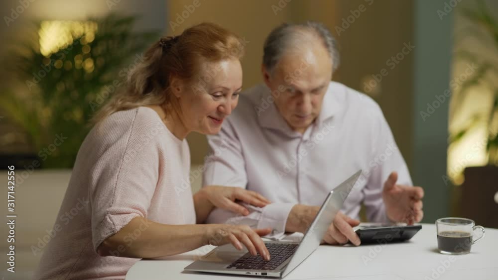 Senior couple sitting at table at home. Wife communicating on laptop computer and showing messages to husband. Husband getting angry and shouting that he is busy calculating home finances Senior couple sitting at table at home. Wife communicating on laptop computer and showing messages to husband. Husband getting angry and shouting that he is busy calculating home finances