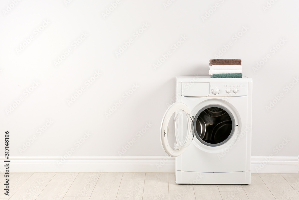Modern washing machine with stack of towels near white wall, space for text. Laundry day