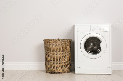 Modern washing machine with laundry basket near white wall