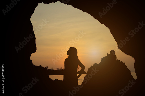 Silhouette of a young woman standing in front of a cave on the beach at sunset