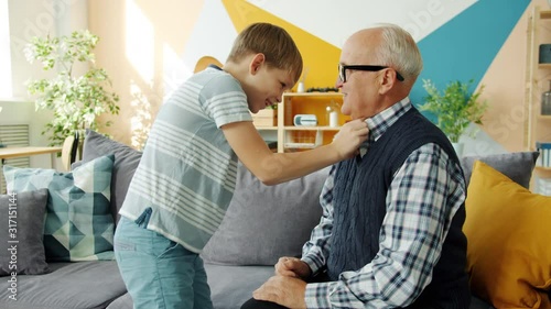 Helpful grandson helping enderly grandfather with clothing while old man is having fun playing with kid at home. Caring people, lifestyle and family concept. photo